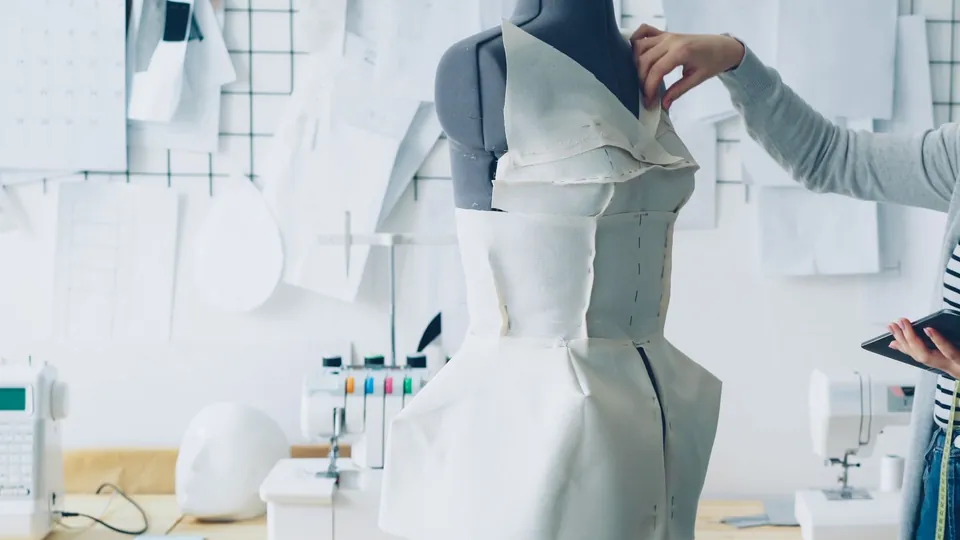 Designer working on a dress on a mannequin in a bright studio, with paper patterns on the wall